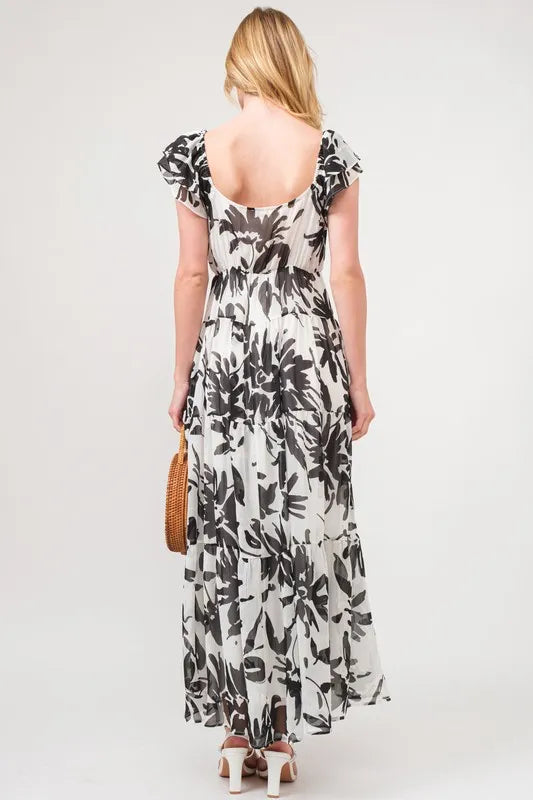 Back view of a woman wearing a black and white floral print tiered maxi dress with ruffled sleeves, a scooped back neckline, and white heeled sandals.