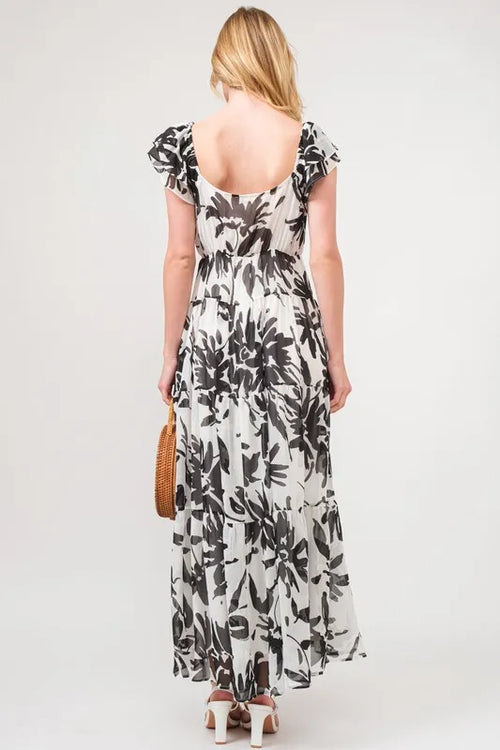Back view of a woman wearing a black and white floral print tiered maxi dress with ruffled sleeves, a scooped back neckline, and white heeled sandals.