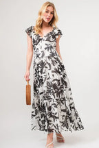 Woman wearing a black and white floral print tiered maxi dress with ruffled sleeves, carrying a woven handbag, and styled with strappy sandals.