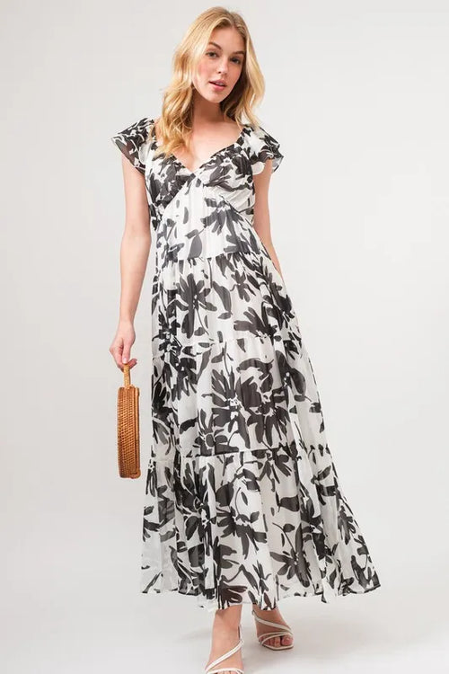 Woman wearing a black and white floral print tiered maxi dress with ruffled sleeves, carrying a woven handbag, and styled with strappy sandals.